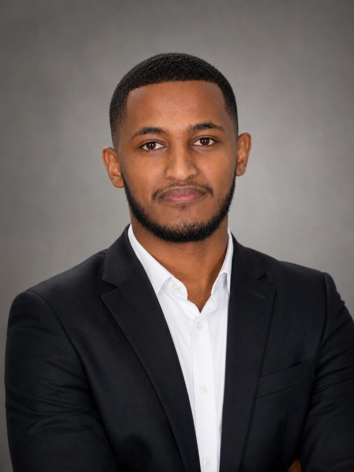 Mukerem Shifa, head and shoulders, in a dark blazer over an open-collar white shirt against a grey studio backdrop.
